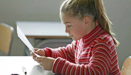 Schülerin, ca. 8 Jahre, Profilbild, Pferdeschwanz und rot geringelter Pulli, liest konzentrirt im Klassenraum von einem Blatt