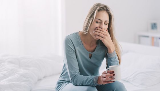 Junge Frau, sitzt mit Kaffee auf dem Bett und gähnt.