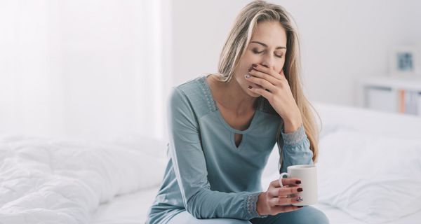 Junge Frau, sitzt mit Kaffee auf dem Bett und gähnt.