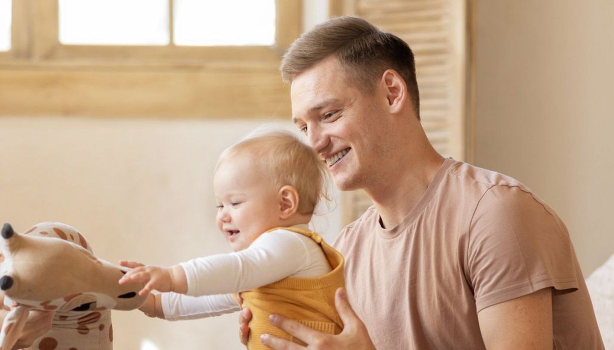 Junge Familie: Der Vater hält das etwa 10 Monate alte Kind, das versucht auf seinen Füßen zu stehen und nach einem Kuscheltier greift.