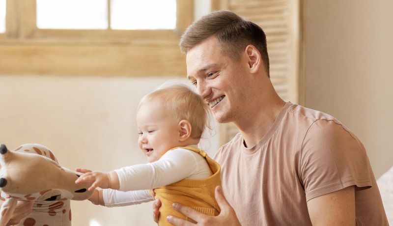 Junge Familie: Der Vater hält das etwa 10 Monate alte Kind, das versucht auf seinen Füßen zu stehen und nach einem Kuscheltier greift.