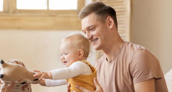 Junge Familie: Der Vater hält das etwa 10 Monate alte Kind, das versucht auf seinen Füßen zu stehen und nach einem Kuscheltier greift.