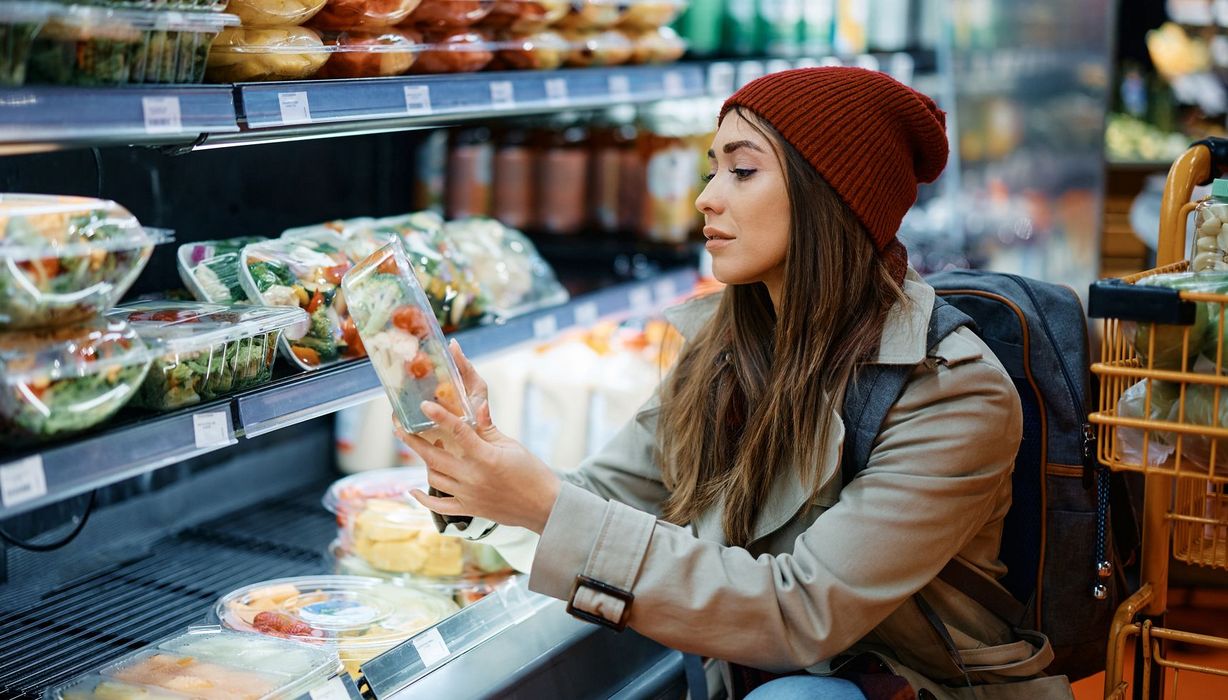 Junge Frau blickt prüfend auf einen Fertigsalat aus dem Kühlregal.