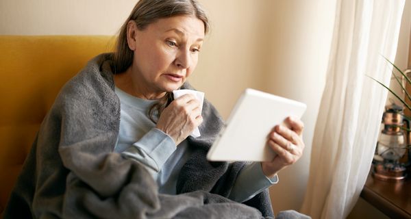 Eine ältere Frau, eingehüllt in einer Decke und mit einem Taschentuch in der Hand, blickt auf ein Tablet.