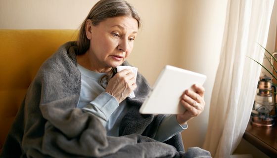 Eine ältere Frau, eingehüllt in einer Decke und mit einem Taschentuch in der Hand, blickt auf ein Tablet. 