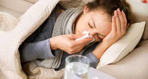 Junge, grippekranke Frau auf der Couch mit Schnupfennase, Hand an der Stirn und Medikamenten 