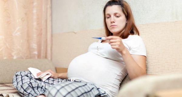 Schwangere Frau sitzt im Bett und schaut auf ein Fieberthermometer