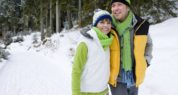 Jüngeres in Kamera lachendes Paar beim Winterspaziergang in grün-gelb-grauen Freizeitklamotten, Mützen, Schals, Mann hat Arm um Frau gelegt, Schneelandschaft 