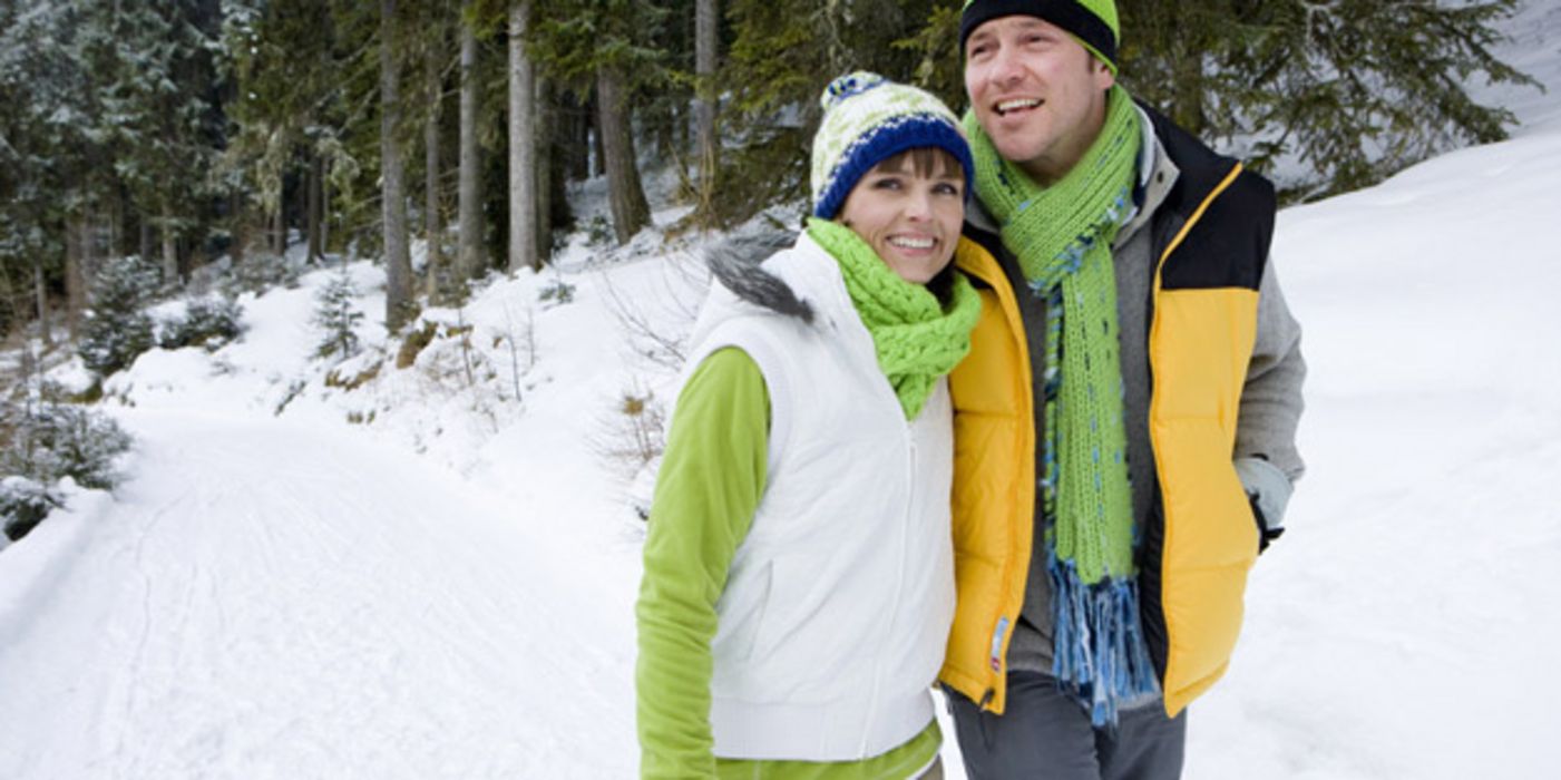 Jüngeres in Kamera lachendes Paar beim Winterspaziergang in grün-gelb-grauen Freizeitklamotten, Mützen, Schals, Mann hat Arm um Frau gelegt, Schneelandschaft 