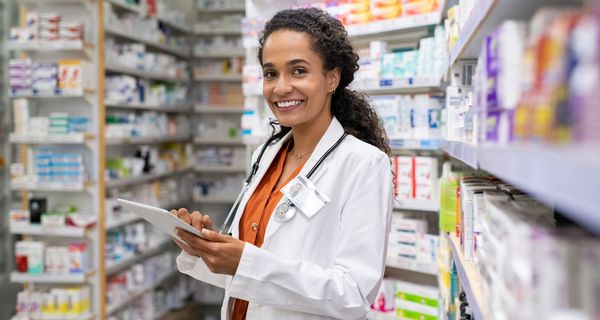 Freundliche Apothekerin, steht mit Tablet hinter einer großen Wand mit Medikamenten.