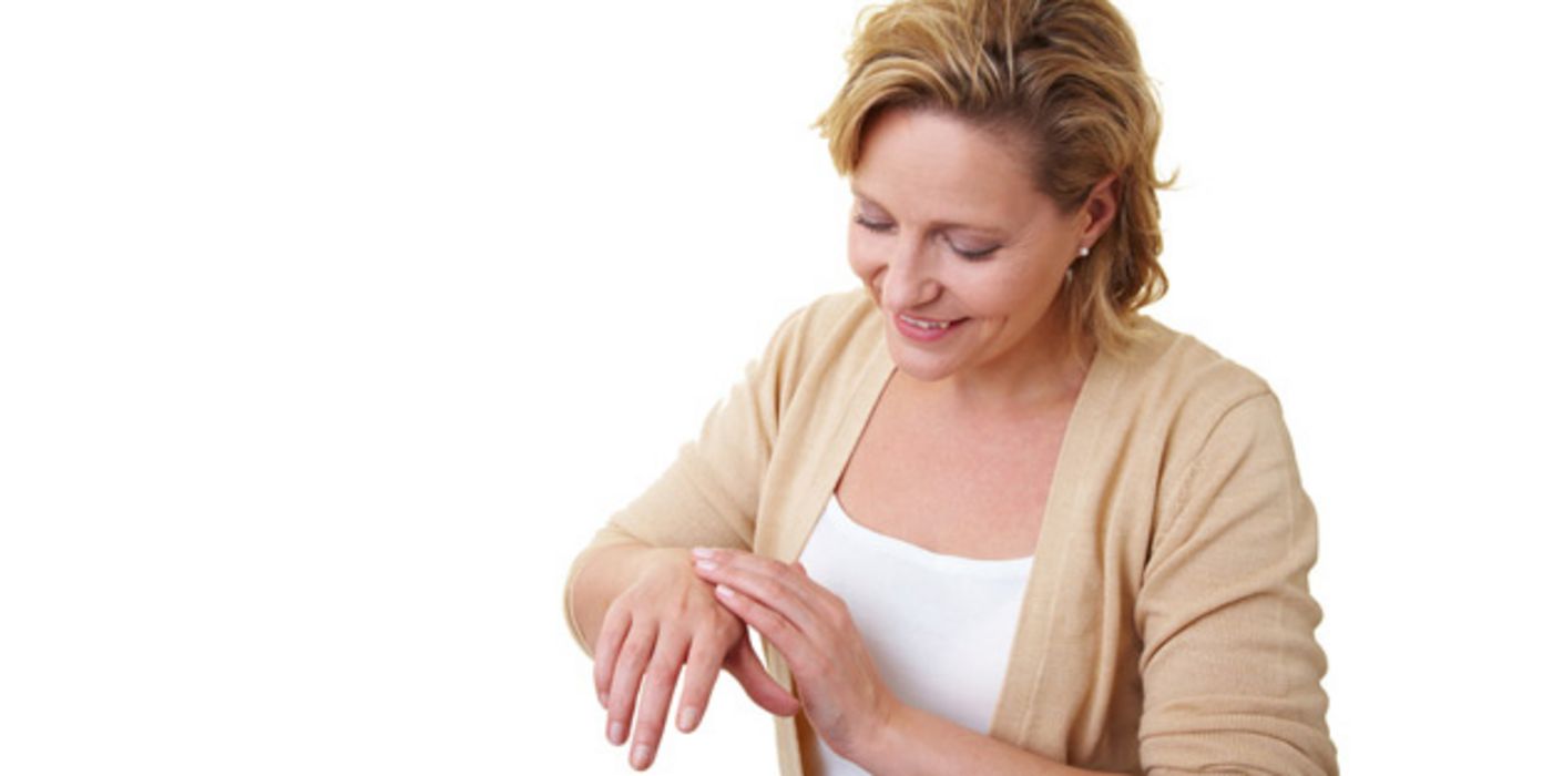 Frau ca. Mitte 40, blond, lächelnd, beim Eincremen der rechten mit der linken Hand. Weißes Top, darüber beige, offene Strickjacke
