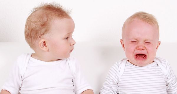 Zwei Zwillings-Babys sitzen nebeneinander: Ein Zwilling weint, der andere schaut ihn verdutzt an.