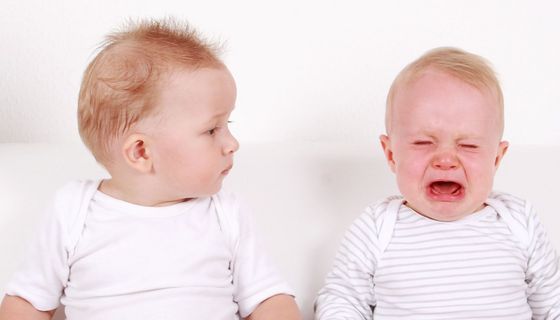 Zwei Zwillings-Babys sitzen nebeneinander: Ein Zwilling weint, der andere schaut ihn verdutzt an.