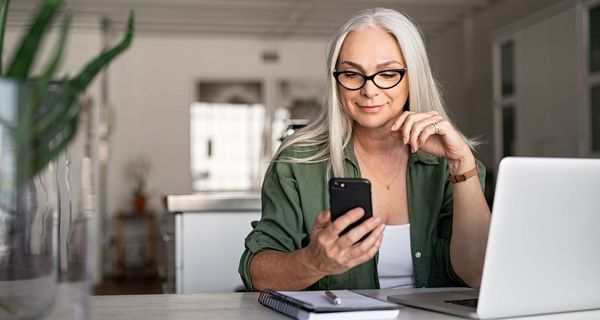 Ältere Frau, ca. 60 Jahre alt, sitzt am Schreibtisch vor einem Laptop mit einem Telefon in der Hand.