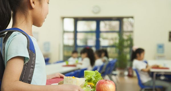 Mädchen trägt ein Tablett mit gesundem Mittagessen – Apfel und Salat sind zu erkennen – zu einem Tisch in der Mensa. Unscharf sind andere Kinder im Hintergrund an den Tischen mit Tabletts zu erkennen.