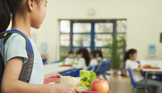 Mädchen trägt ein Tablett mit gesundem Mittagessen – Apfel und Salat sind zu erkennen – zu einem Tisch in der Mensa. Unscharf sind andere Kinder im Hintergrund an den Tischen mit Tabletts zu erkennen.