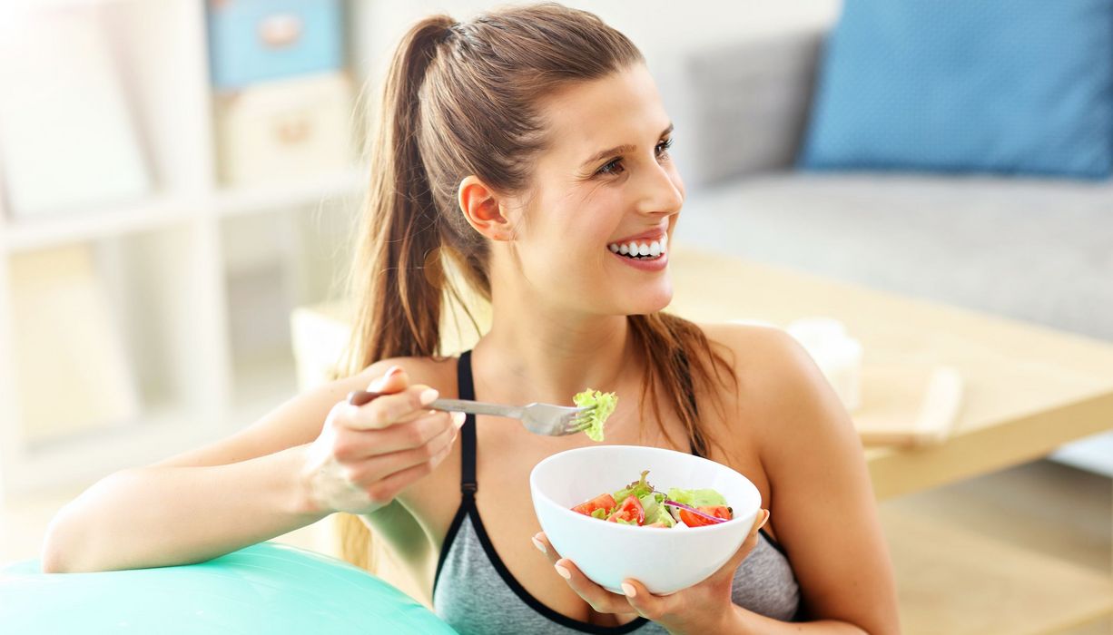 Frau isst in Sportkleidung einen Salat und lehnt auf einem Gymnastikball. 
