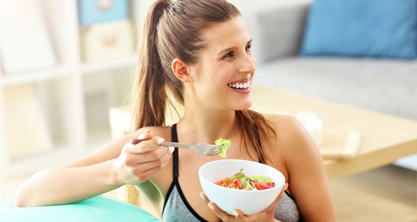 Frau isst in Sportkleidung einen Salat und lehnt auf einem Gymnastikball. 