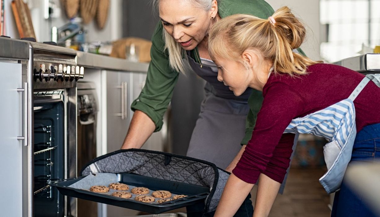 Eine ältere Frau nimmt mithilfe eines Hitzeschutzes ein heißes Blech mit gebackenen Keksen aus einem Ofen. Ein Mädchen hält die Ofentür offen - ihre nackten Unterarme kommen dabei dem Blech gefährlich nahe.