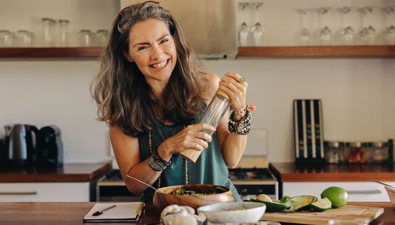 Eine Frau Ü50 pfeffert ihr essen mit einer Mühle und lächelt in die Kamera.