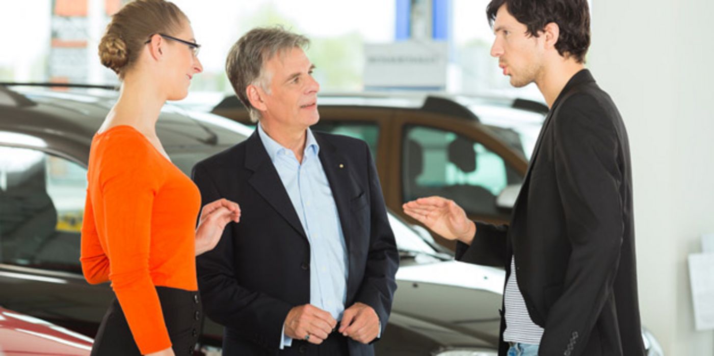 Junges Paar in Verhandlung mit einem älteren Autoverkäufer, Autos im Hintergrund
