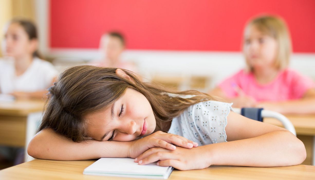 Ein müdes Mädchen hat den Kopf auf die Arme und den Tisch in der Schulklasse gelegt. Im Hintergrund sind unscharf andere Schulkinder zu sehen. 