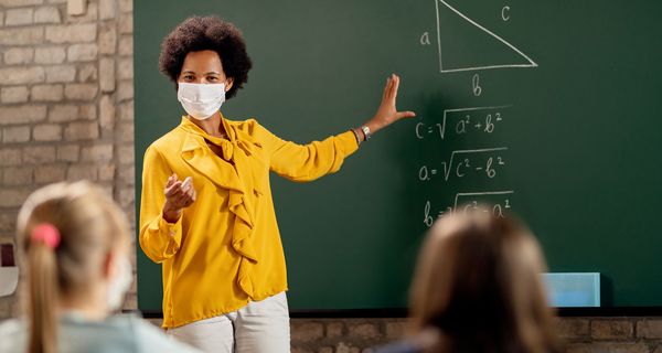 Lehrerin mit Maske steht an der Tafel und erklärt ihren Schülern eine Matheaufgabe.