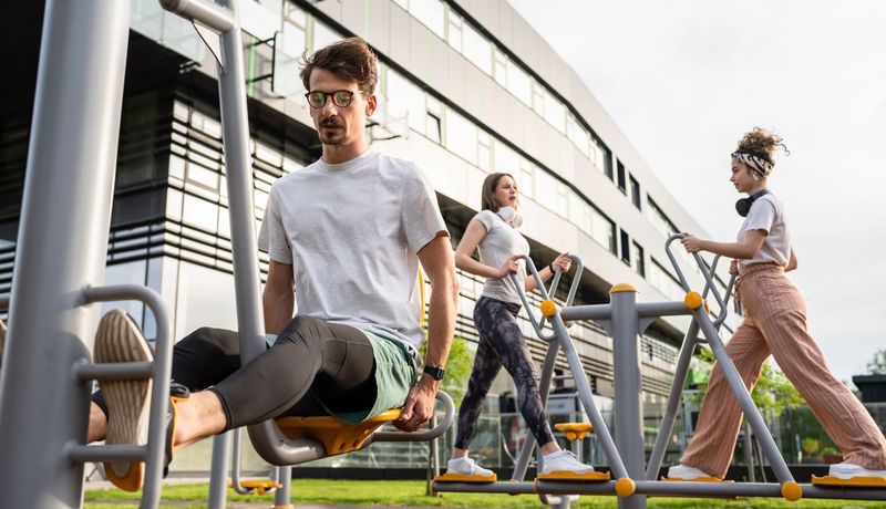Ein Mann trainiert im Sitzen an einem Outdoor-Gerät, im Hintergrund sind zwei Frauen in Schrittstellung, ebenfalls auf Geräten,  zu sehen. 