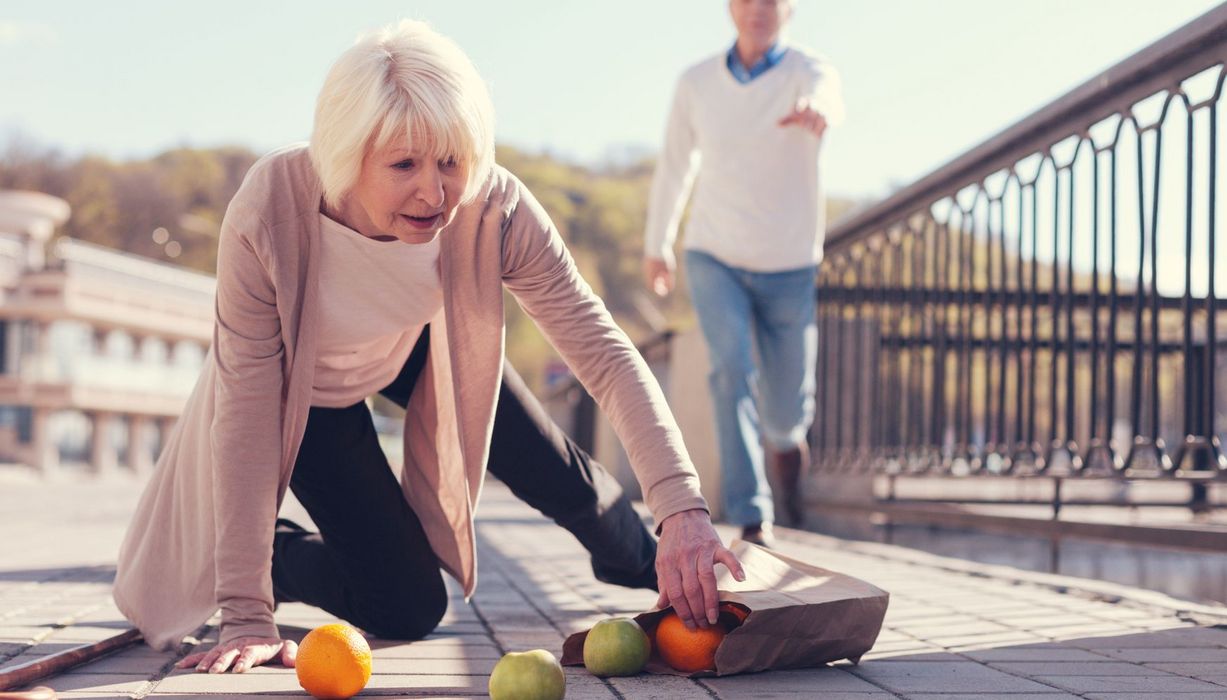 Eine Frau über 65 ist gestürzt, sie sammelt bereits Obst wieder ein. Im Hintergrund sieht man unscharf jemanden, der ihr zur Hilfe eilen will.