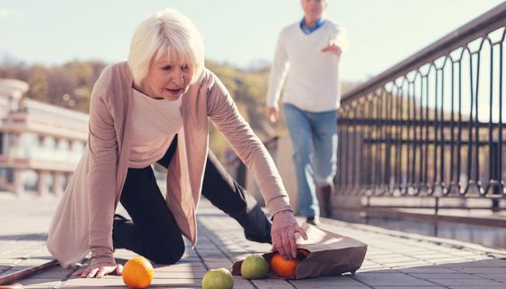 Eine Frau über 65 ist gestürzt, sie sammelt bereits Obst wieder ein. Im Hintergrund sieht man unscharf jemanden, der ihr zur Hilfe eilen will.