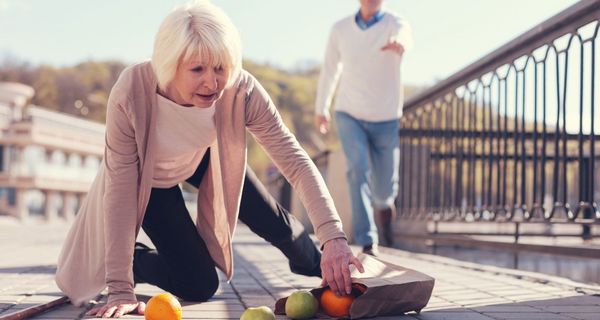 Eine Frau über 65 ist gestürzt, sie sammelt bereits Obst wieder ein. Im Hintergrund sieht man unscharf jemanden, der ihr zur Hilfe eilen will.