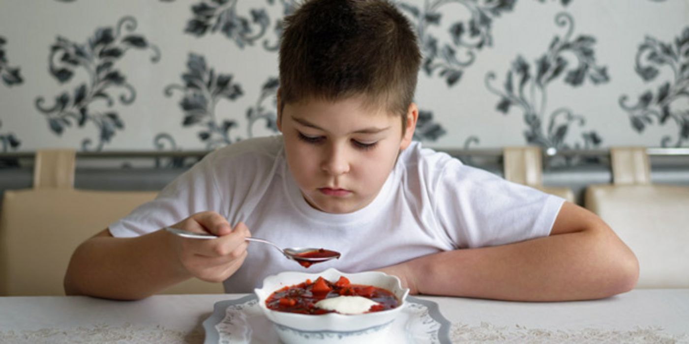Junge sitzt am Esstisch vor einem Teller Suppe und hält einen Löffel in der Hand, den er kritisch beäugt 