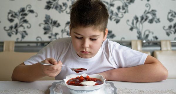 Junge sitzt am Esstisch vor einem Teller Suppe und hält einen Löffel in der Hand, den er kritisch beäugt 