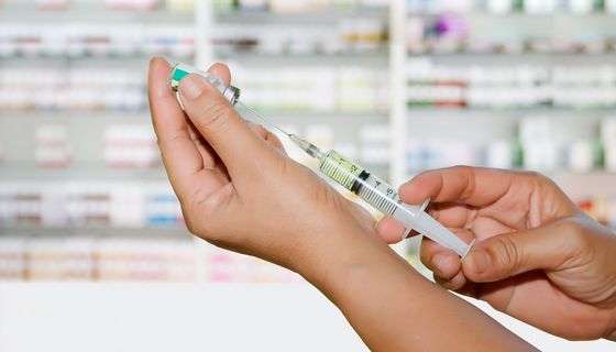 Hand, hält eine Spritze, im Hintergrund eine Apotheke.