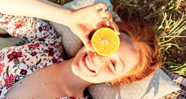 Junge Frau, liegt auf einer Wiese und hält seine Orange vor ein Auge.