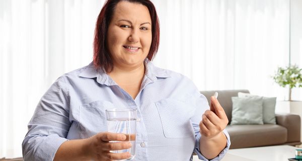 Eine Frau mit Übergewicht hält lächelnd eine Tablette und ein Glas Wasser in der Hand.