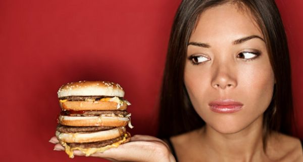 Junge Frau schaut sehr skeptisch auf einen mehrstöckigen Cheeseburger auf ihrer Handfläche