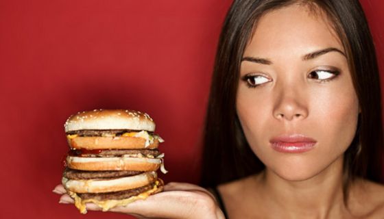 Junge Frau schaut sehr skeptisch auf einen mehrstöckigen Cheeseburger auf ihrer Handfläche