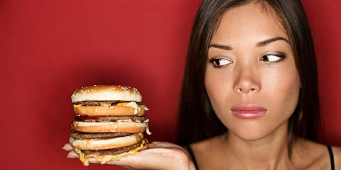 Junge Frau schaut sehr skeptisch auf einen mehrstöckigen Cheeseburger auf ihrer Handfläche