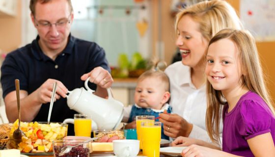 Mutter, Vater, Tochter (ca. 8 Jahre alt) und Baby beim vergnügten Frühstück, blonde Tochter im Vordergrund lächelt in die Kamera
