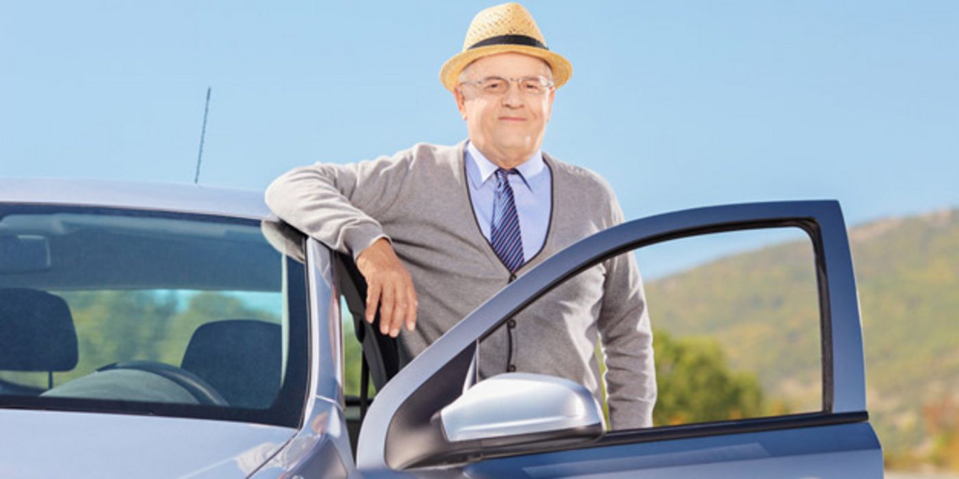 Sommerbild draußen (blauer Himmel, grüne Hügel): Älterer Mann, Strohhut, leichter grauer Sweater, hellblaues Hemd, dunkelblauer Schlips mit weißen diagonalen Streifen, lehnt lächelnd an Autodach, offene Fahrertür, er dahinter