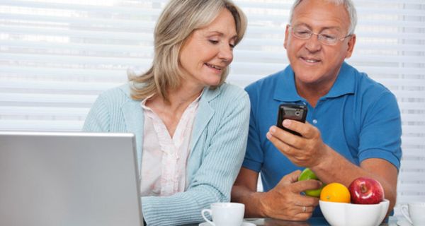 Fittes, attraktives Paar (Frau, längere graue Haare, Mann gebräunt, graue Haare), am Tisch, Laptop links im Bild, schauen gemeinsam auf Smartphone, das Mann in der linken Hand hält