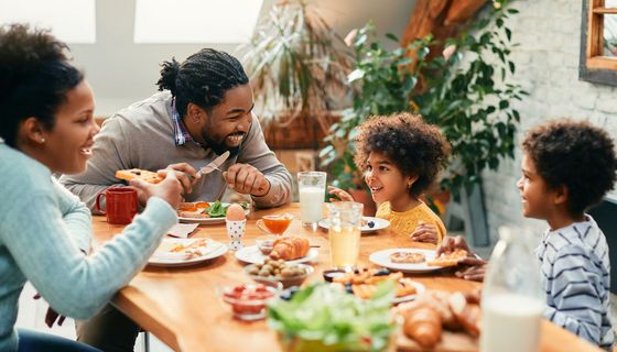 Eine Familie – Eltern mit zwei Kindern – am gedeckten Tisch mit Salat, Saft, Tomaten, Oliven und Spiegelei.