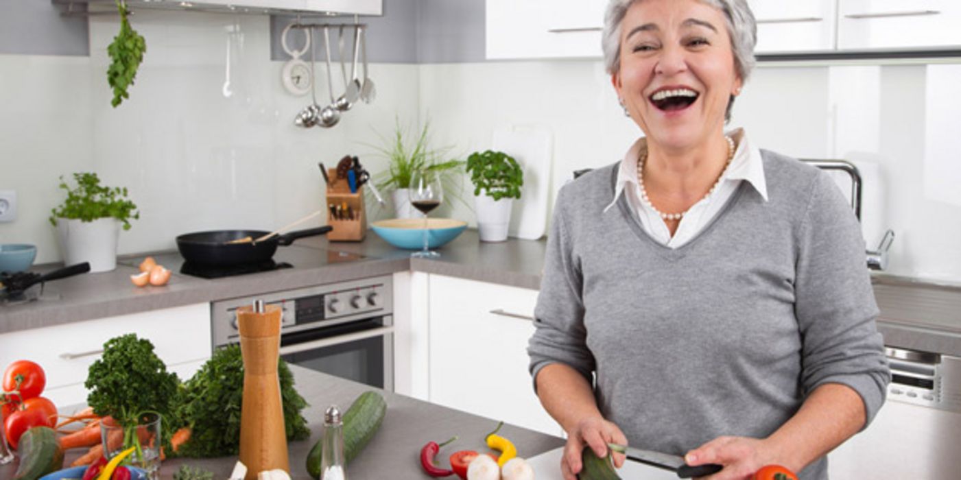 Lachende Frau mit kurzen grauen Haaren beim Gemüseschneiden in der Küche
