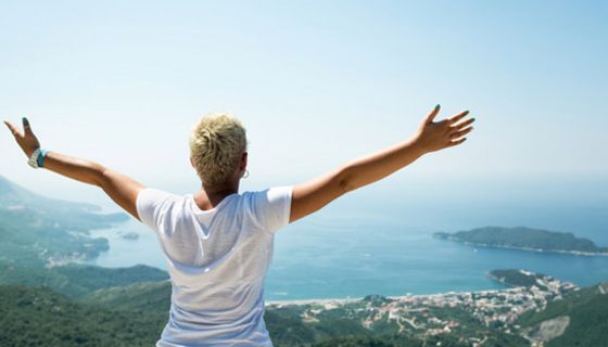Rückenansicht: Frau, kurze, blonde Haare, weiße Bluse, auf Berg stehend auf Meer und Inseln schauend, ausgebreitete Arme