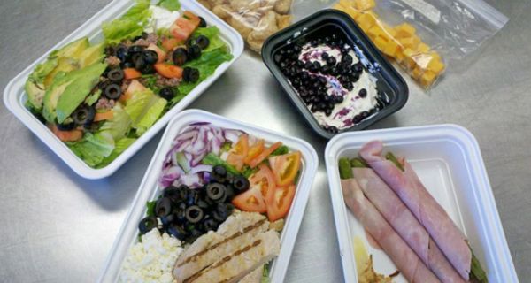 Verschiedene Lebensmittel (Schinkenröllchen, gemischter Salat mit Oliven, Dessert mit Heidelbeeren, Käsewürfel) in Styroporschalen oder Plastiktüten