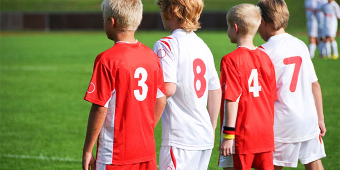 4 Fußballjungs, blond, Rückansicht, etwas Profil, auf Platz, abwechselnd rote Trikots mit weißer Nr. und umgekehrt tragend 