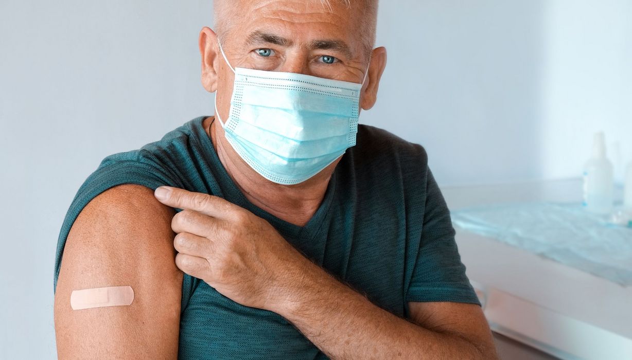 Älterer Mann mit blauer OP-Maske, zeigt seinen Oberarm mit Pflaster von seiner Impfung.