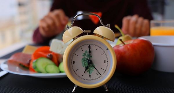 Im Vordergrund eine Uhr: Dahinter eine Person, die isst (nur die Hände sichtbar, sowie ein Apfel, Orangensaft, Gruke, Paprika, Wurst und Käse). 