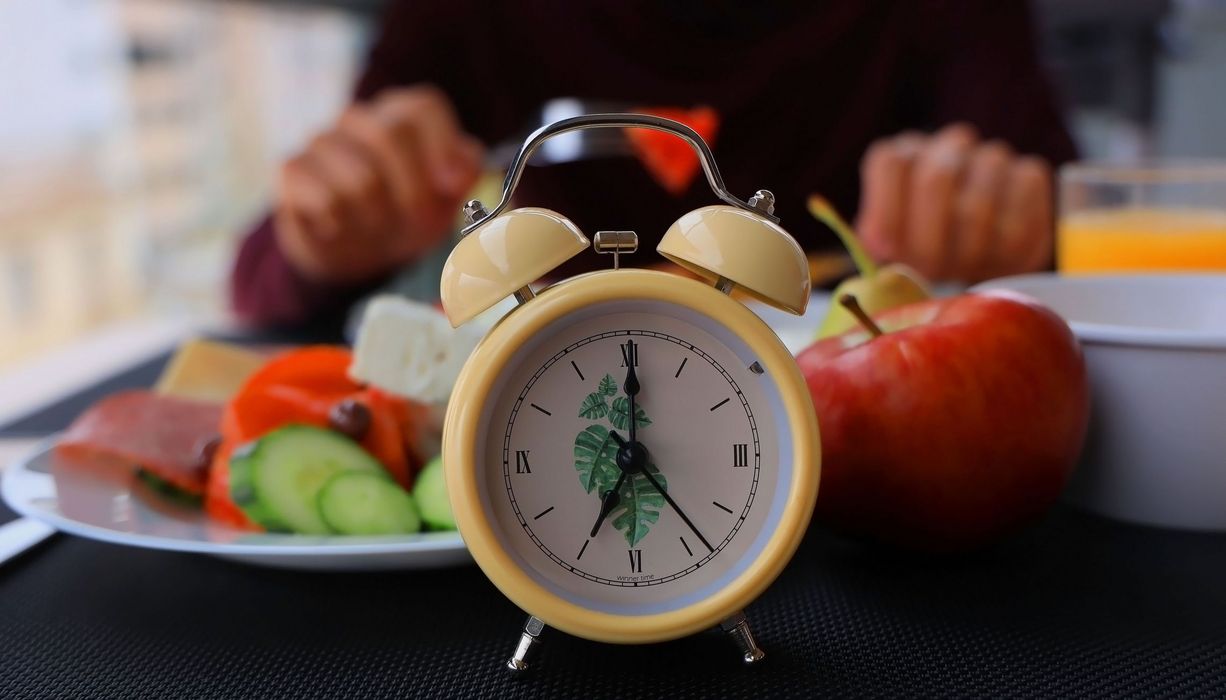 Im Vordergrund eine Uhr: Dahinter eine Person, die isst (nur die Hände sichtbar, sowie ein Apfel, Orangensaft, Gruke, Paprika, Wurst und Käse). 